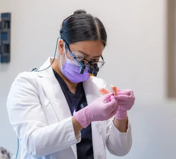 Dentist examining a denture model wearing protective gear and magnification loupes.